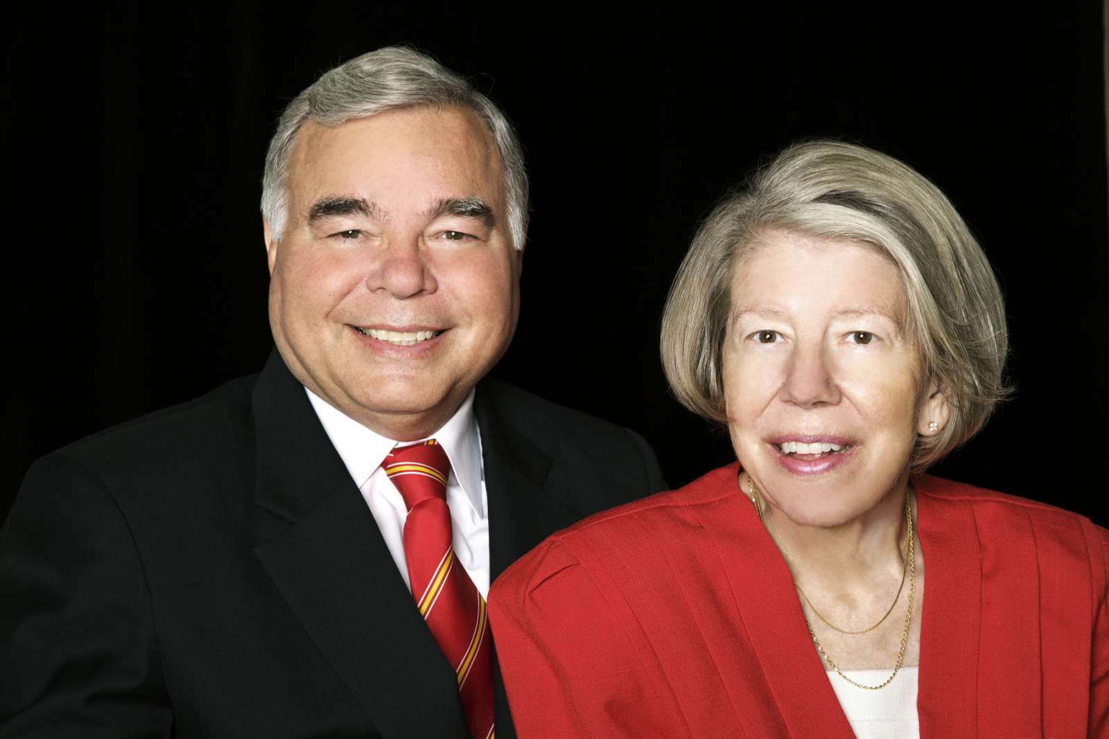 Thomas Smith, Evening Undergraduate Commencement Ceremony Speaker, stands with his wife, Evonne. He is wearing a black suit jacket, white shirt, and a red and gold striped tie. She is wearing a red suit jacket, white shirt, and a necklace.